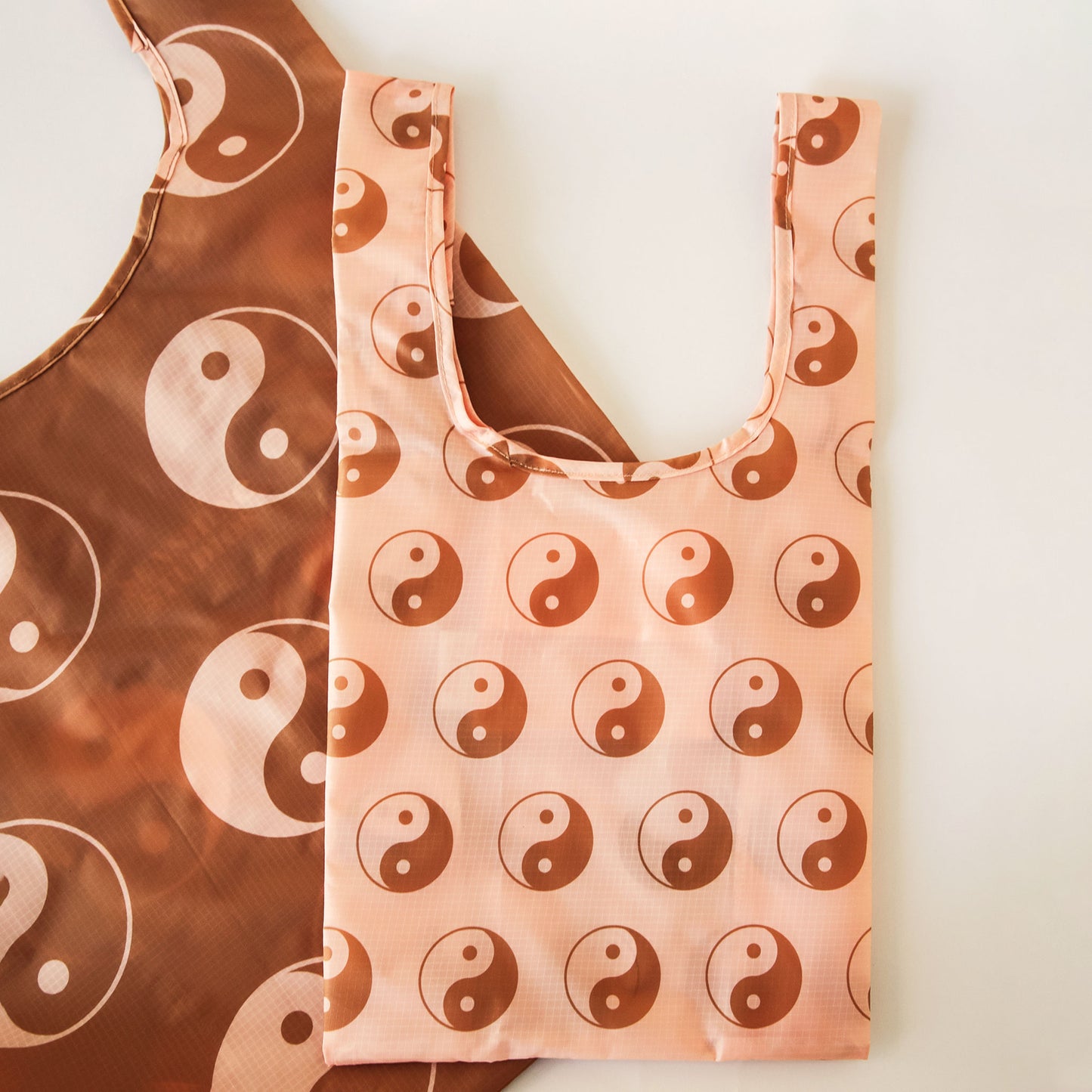 Peach reusable bag covered in a print of brown and peach yin-yang symbols. The bag is positioned flat on a table and has a 'U' shape between two handles. A larger brown bag lays behind and is covered in larger peach and brown yin-yang symbols. 