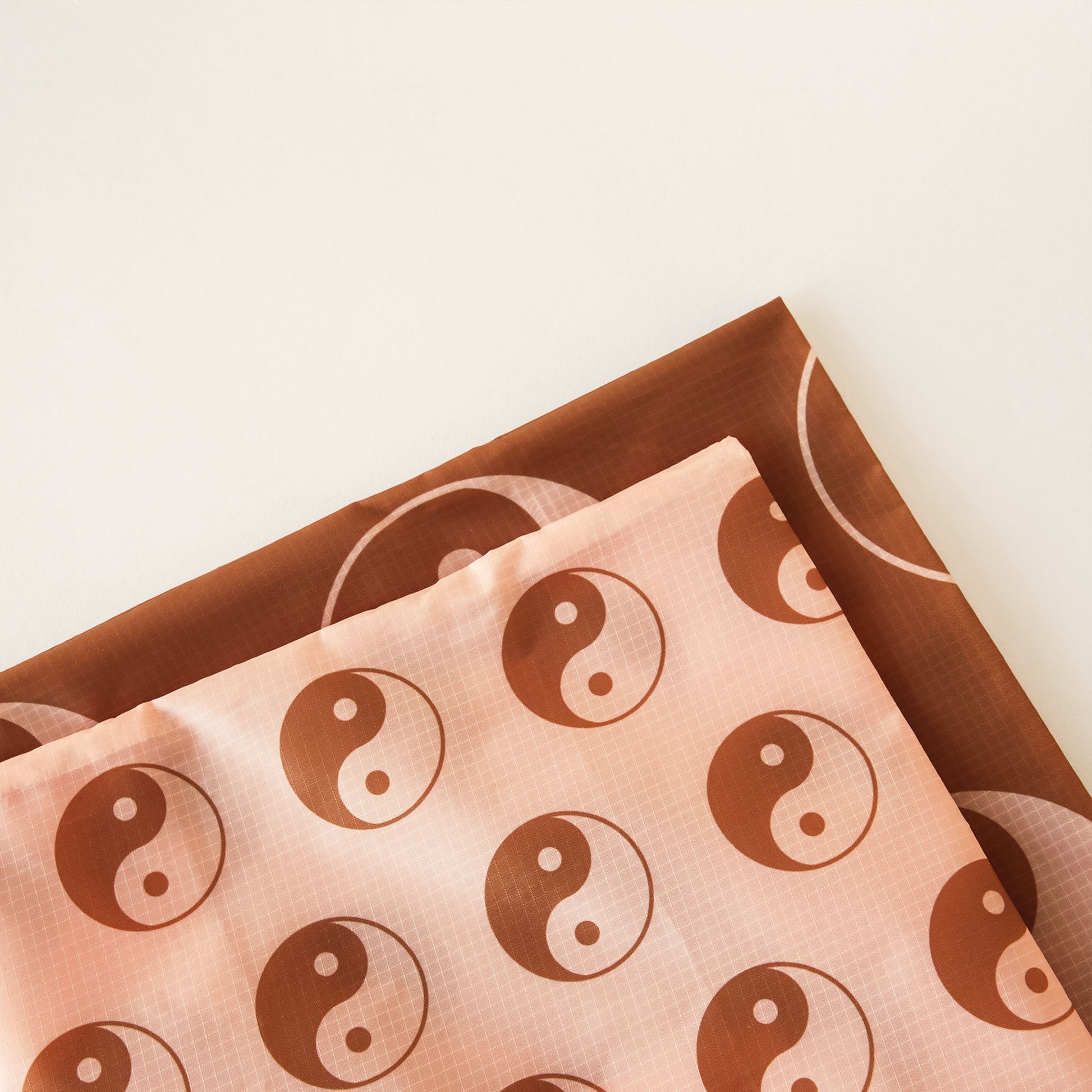 Zoomed in view of the corner of two bags. The bag layered above is peach and covered in brown and peach yin-yang symbols. The bag below is brown with larger yin-yang symbols. 