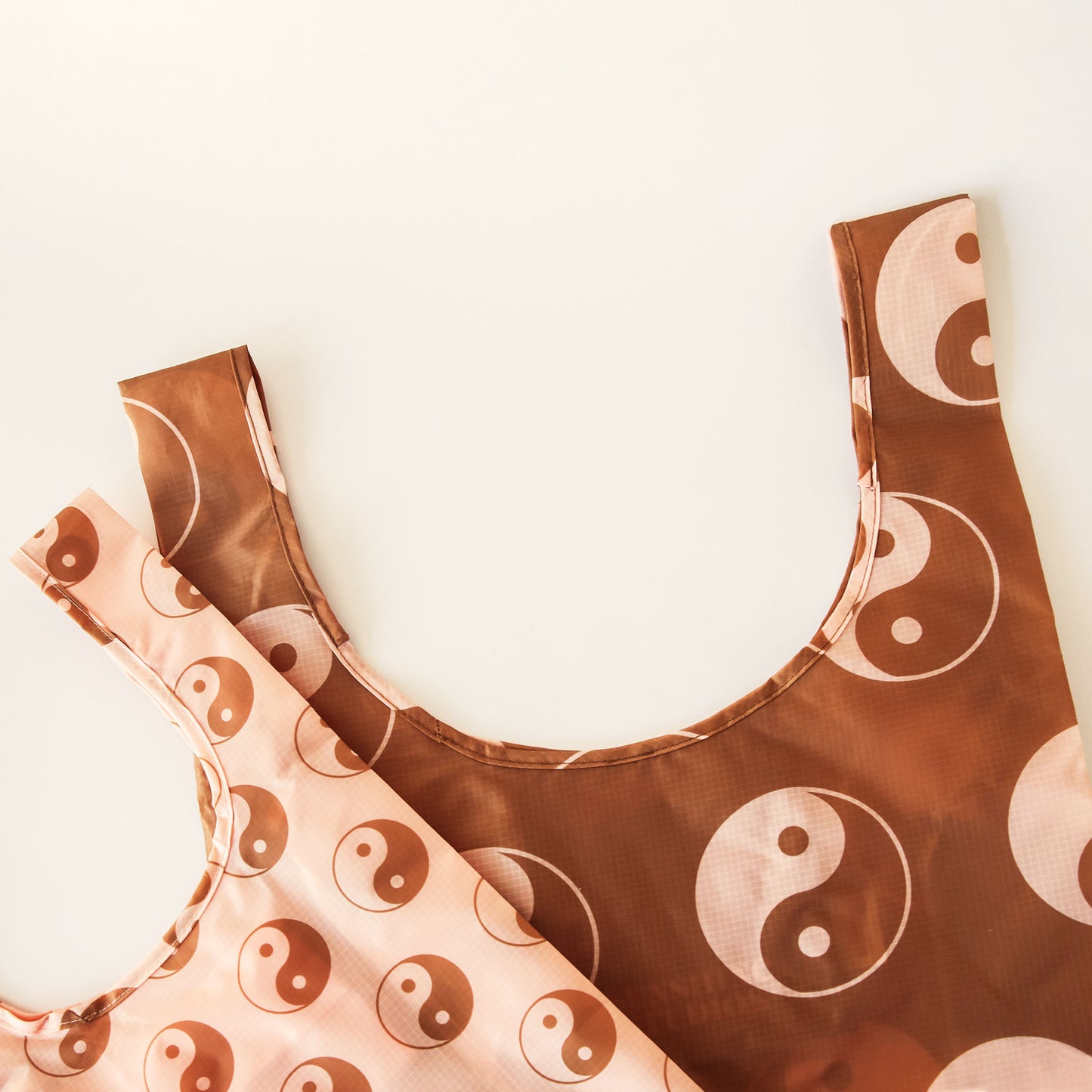 Zoomed in view of two nylon reusable bags. The bag to the left is brown with peach and brown yin-yang symbols. The bag the left is peach with smaller brown and peach yin-yang symbols.