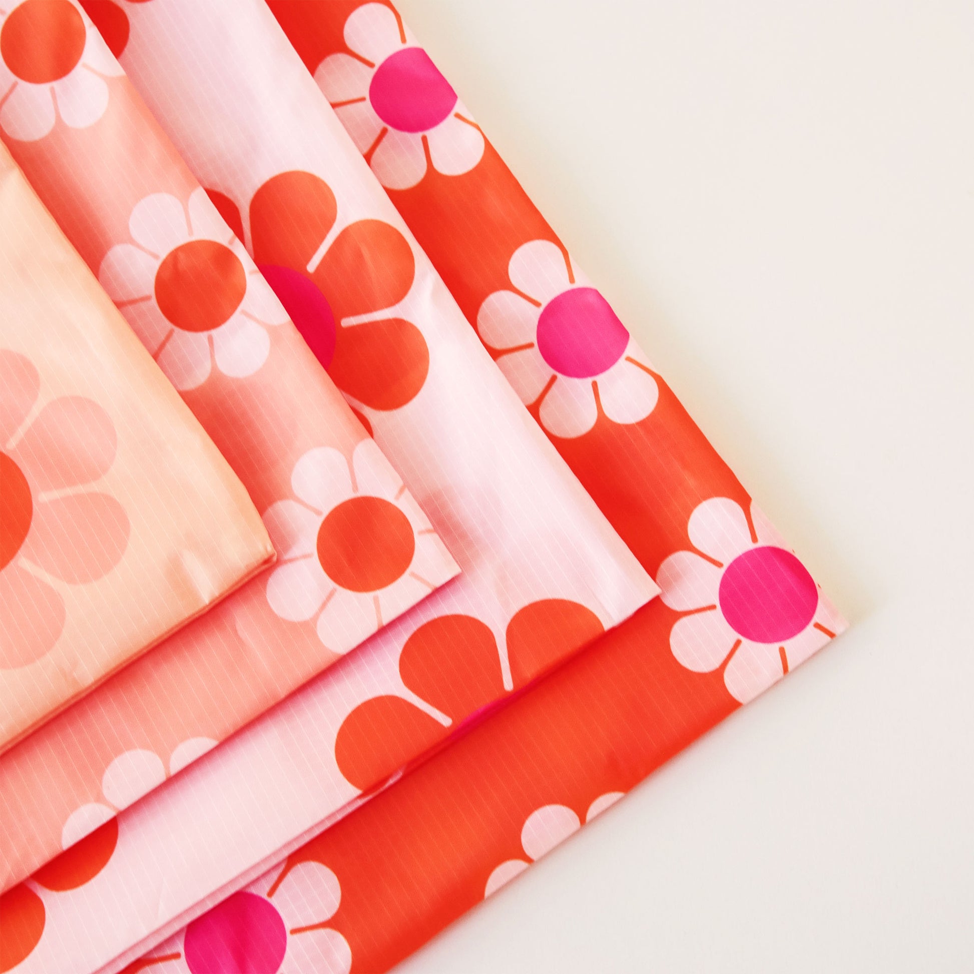 Zoomed in view of the corners of four reusable bags. Each bag has similar print of daisies but in different combinations of peach, orange and pink.