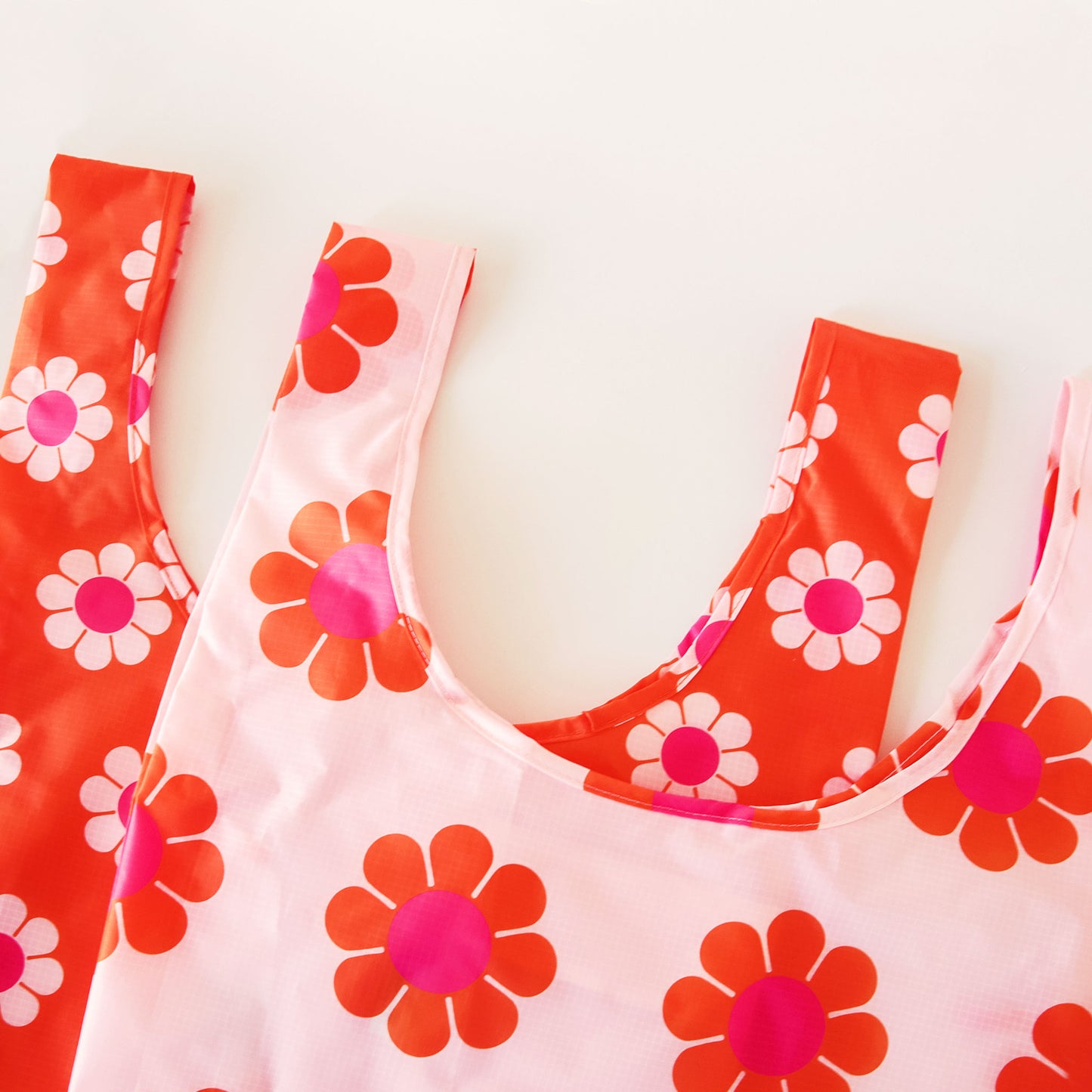 Two nylon bags layered on top of each other. Both bags have a similar print of daisy flowers. The bag layered below is red-orange with peach and pink flowers and the bag above is peach with pink and red-orange flowers.