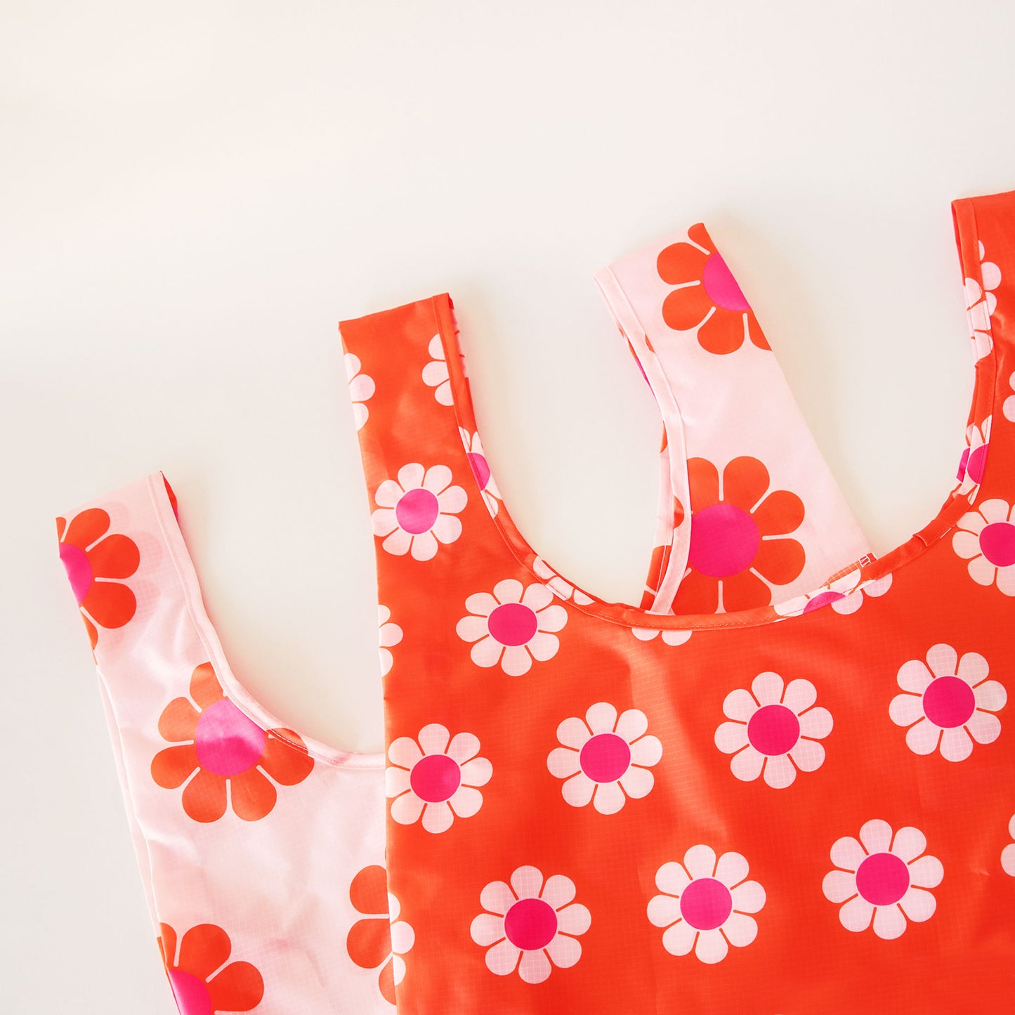 Two nylon bags layers on top of each other. Both bags have a similar print of daisy flowers. The bag layered bellow is peach with orange and pink flowers and the bag above is red-orange with pink and peach flowers 