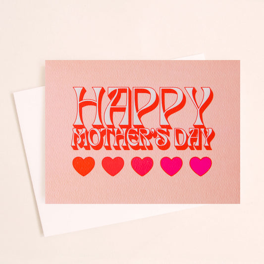 Soft pink card that reads 'Happy Mother's Day' in retro white lettering with a red shadow. Below is a row of five hearts in various reds and pinks. The card is accompanied by a solid white envelope.