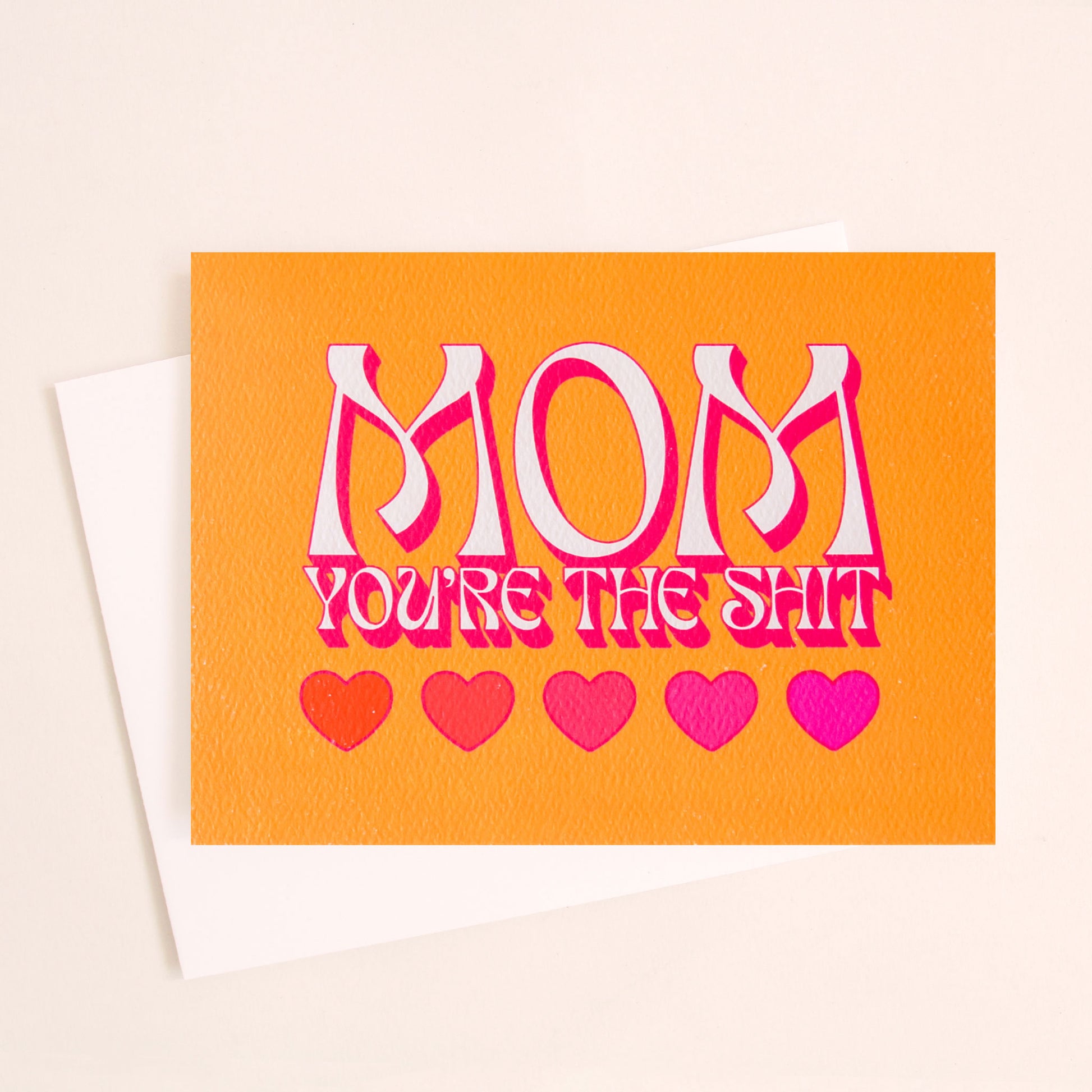 Bright orange card that reads 'Mom You're the Shit' in retro white text with a magenta shadow. Below is a row of five hearts in various reds and pinks. The card is accompanied by a solid white envelope.