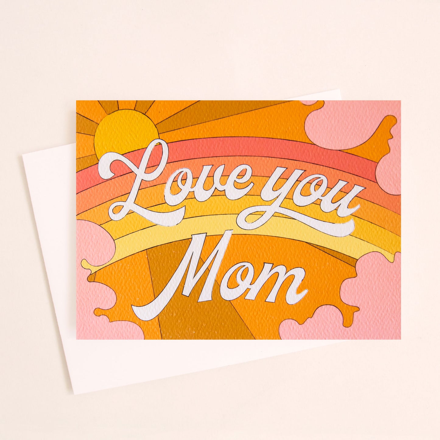 Card with a vibrant scene of a warm toned rainbow and beaming yellow sun. Soft pink clouds take up the edges. The center of the card reads 'Love You Mom' in white cursive lettering. 