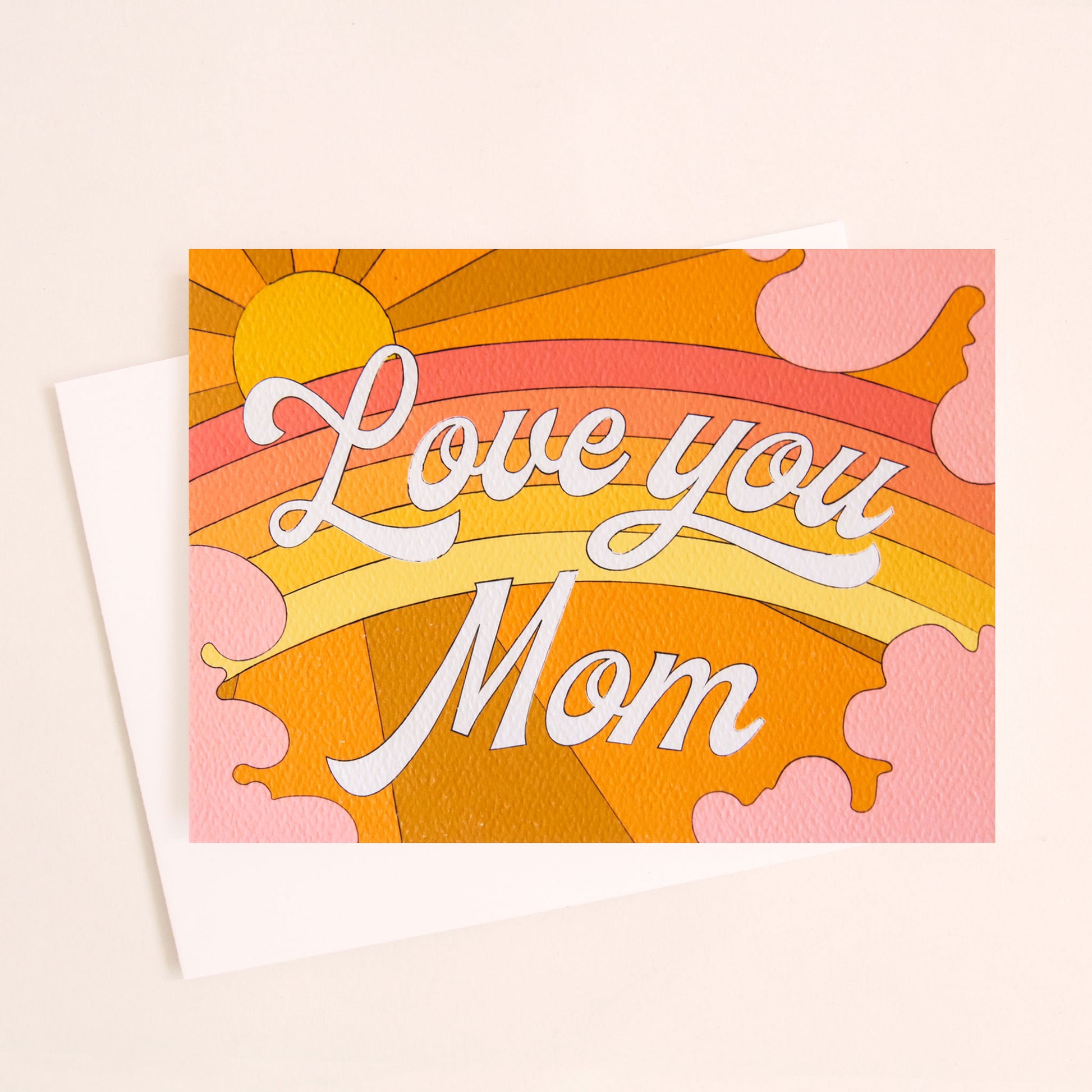 Card with a vibrant scene of a warm toned rainbow and beaming yellow sun. Soft pink clouds take up the edges. The center of the card reads 'Love You Mom' in white cursive lettering. 