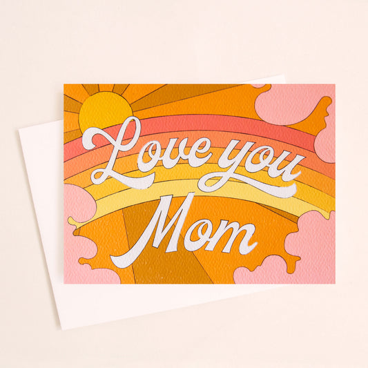 Card with a vibrant scene of a warm toned rainbow and beaming yellow sun. Soft pink clouds take up the edges. The center of the card reads 'Love You Mom' in white cursive lettering. 