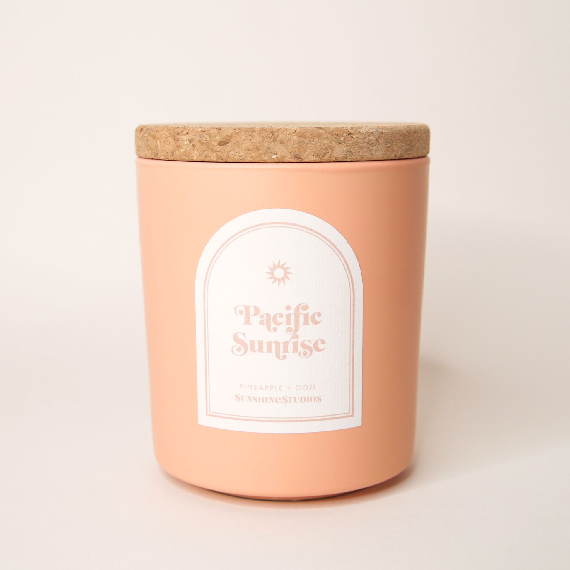 Pastel orange candle with lid made of cork. An arched label reads 'Pacific Sunrise, pineapple + goji, sunshine studios' in soft pink lettering. A small sun design sits above the text.