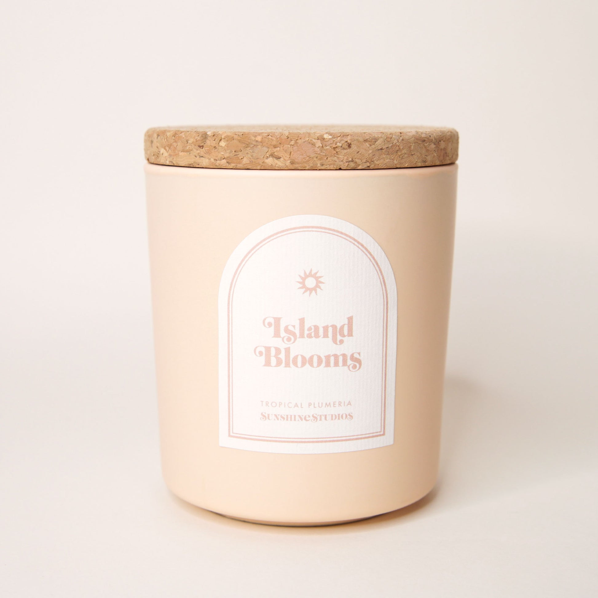 Soft beige candle with lid made of cork. An arched label reads 'Island Blooms, tropical plumeria, sunshine studios' in soft pink lettering. A small sun design sits above the text.
