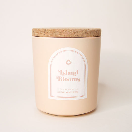 Soft beige candle with lid made of cork. An arched label reads 'Island Blooms, tropical plumeria, sunshine studios' in soft pink lettering. A small sun design sits above the text.