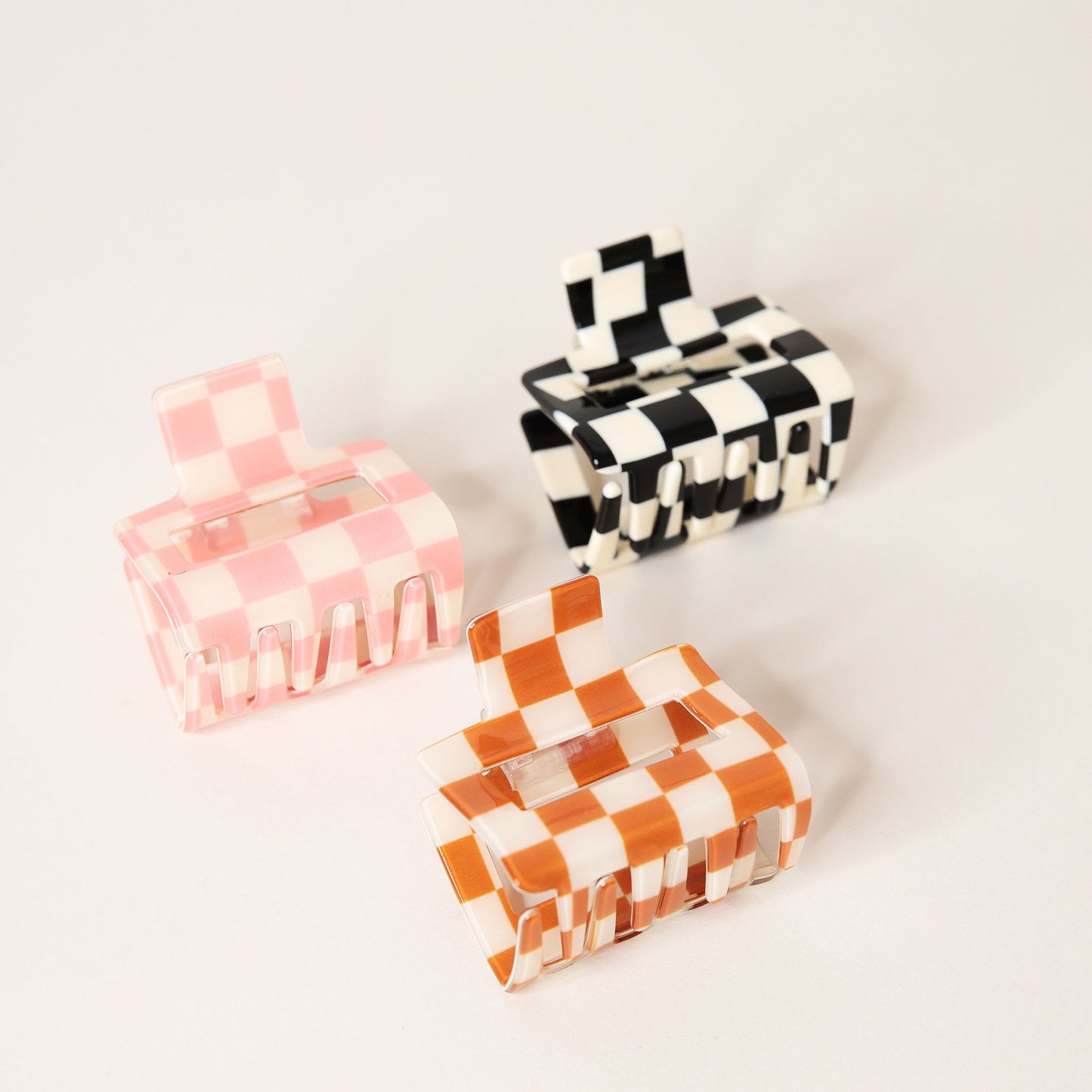 Three checkered claw hair clips in shades of pink, black and orange. Each is a square shape and has two simple geometric cutouts. 