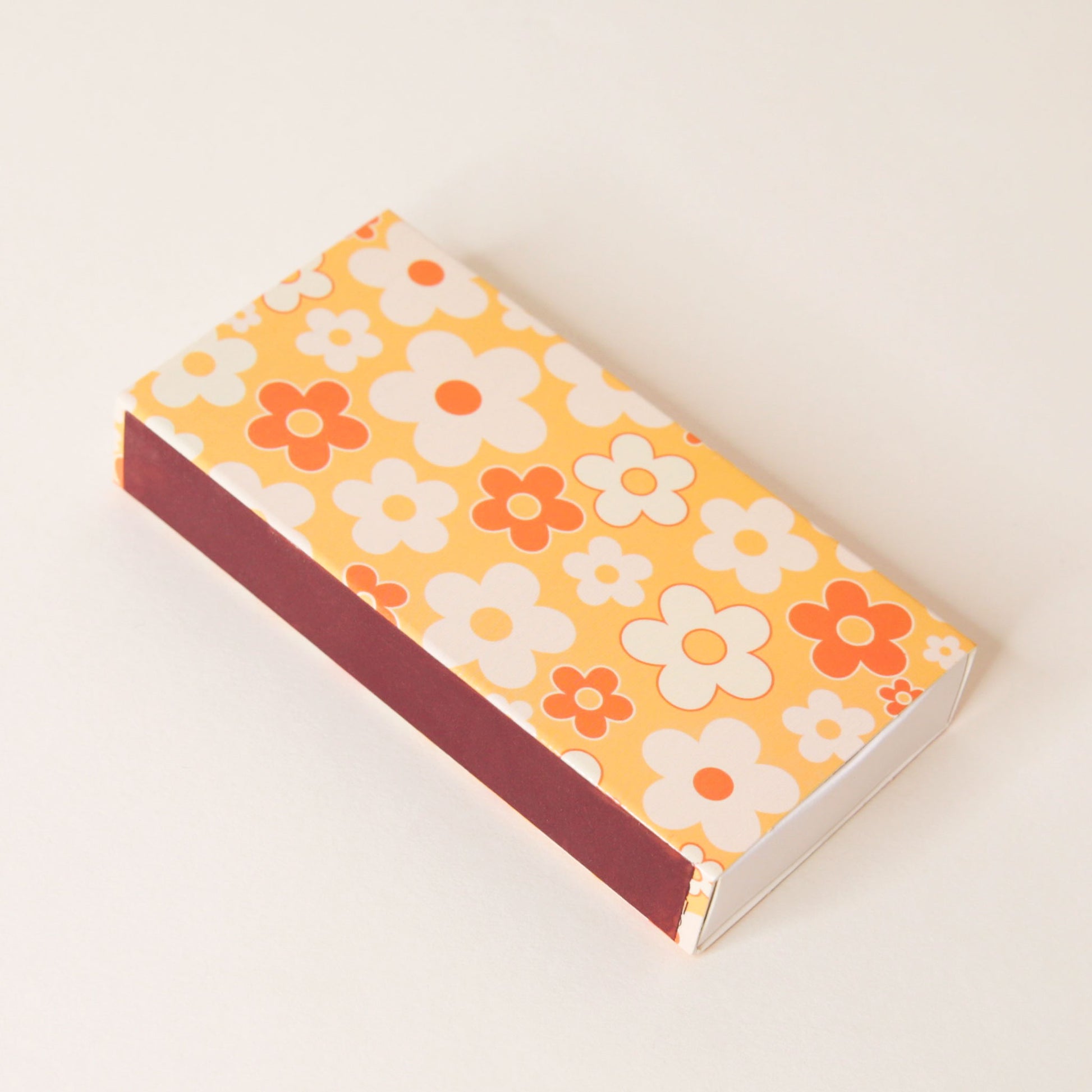 Rectangular match box covered in a flower pattern filled with yellow oranges and whites. The side of the match box is covered in a maroon striking surface.