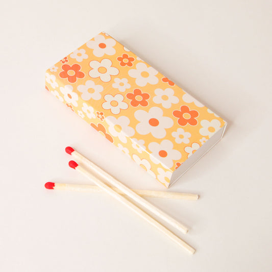 Rectangular match box covered in a flower pattern filled with yellow oranges and whites. Three matches with red dipped tips lay besides the box.