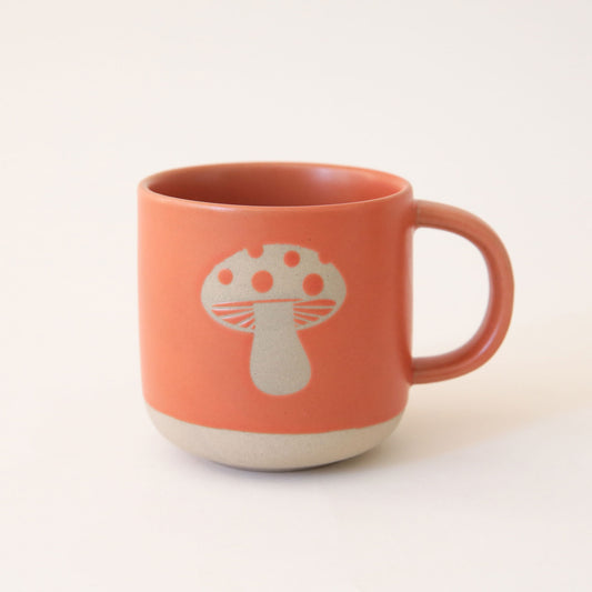 Natural clay mug painted dark orange with silhouette of toad stool mushroom on the front. The base of the mug is unpainted, revealing natural clay.
