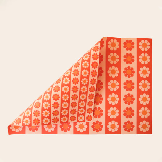 Sheet of wrapping paper filled with stripes of bright and pastel orange flowers. The sheet is bent forward, revealing the other side of the wrapping paper. The back side is covered in thinner stripes of the same floral print. 