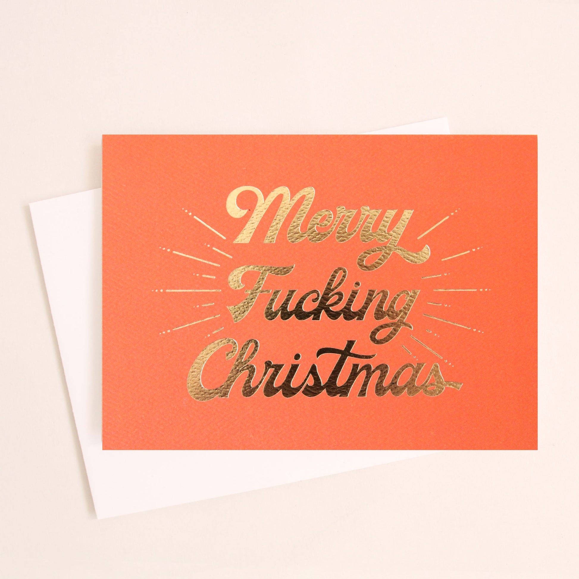 Red orange card that reads 'Merry Fucking Christmas' in cursive gold foil lettering'.  A gold foil burst detailing beams from the text.