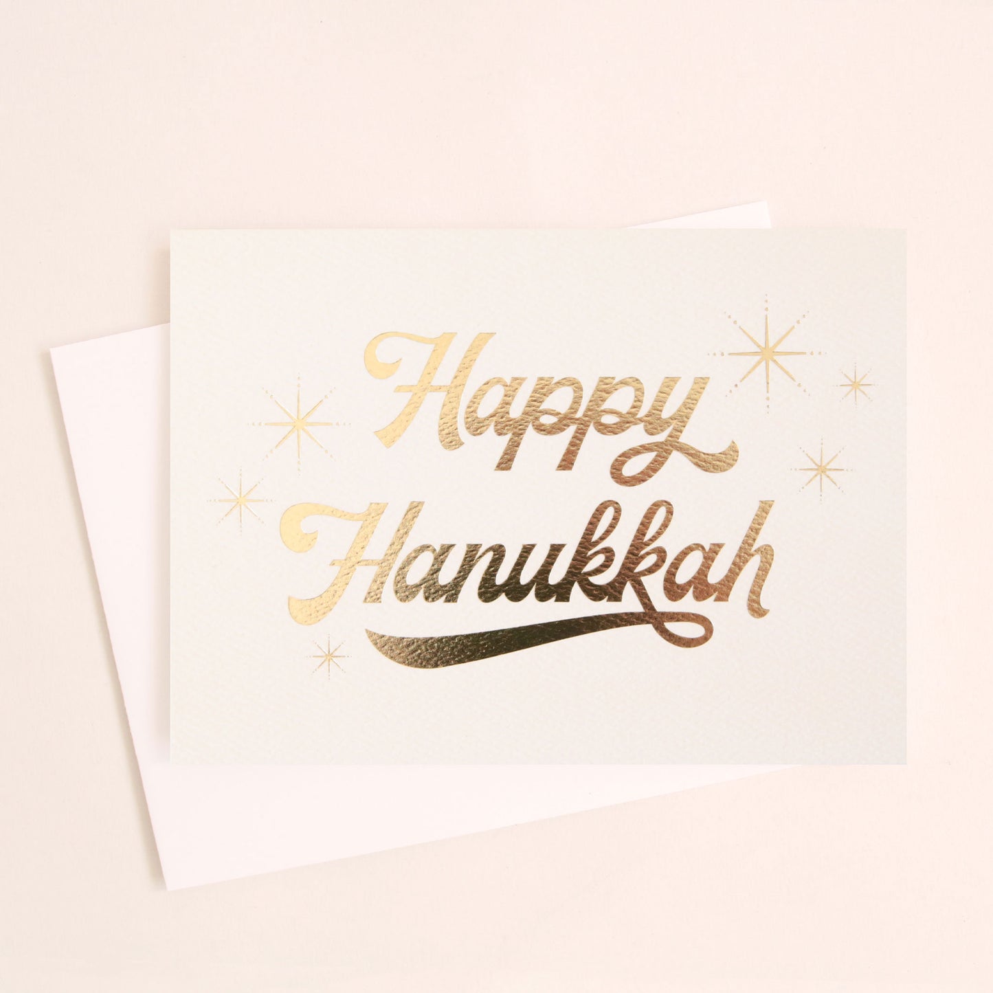 Cream greeting card that reads 'Happy Hanukkah' in gold foil cursive lettering. Gold foil stars twinkle around the text. The card is accompanied by a solid white envelope. 
