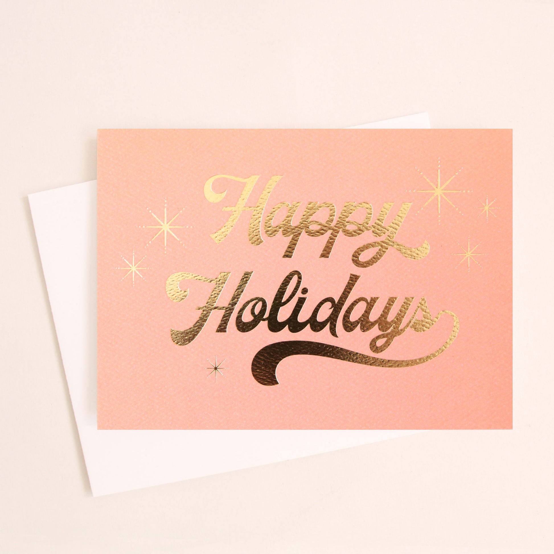 peach colored greeting card with cursive text in gold foil that reads happy holidays surrounded by six gold foil starbursts. The card is accompanied by a solid white envelope. 