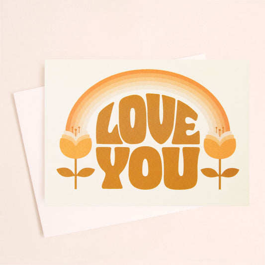 A white card and white envelope with orange 70's text that reads, "Love You". The illustration on the card is an orange toned rainbow feeding into two tulip flowers on either side.