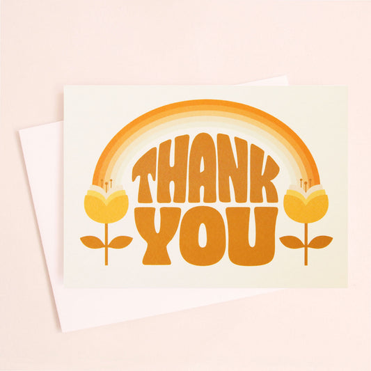 A white card and white envelope with orange 70's text that reads, "Thank You". The illustration on the card is an orange toned rainbow feeding into two tulip flowers on either side.