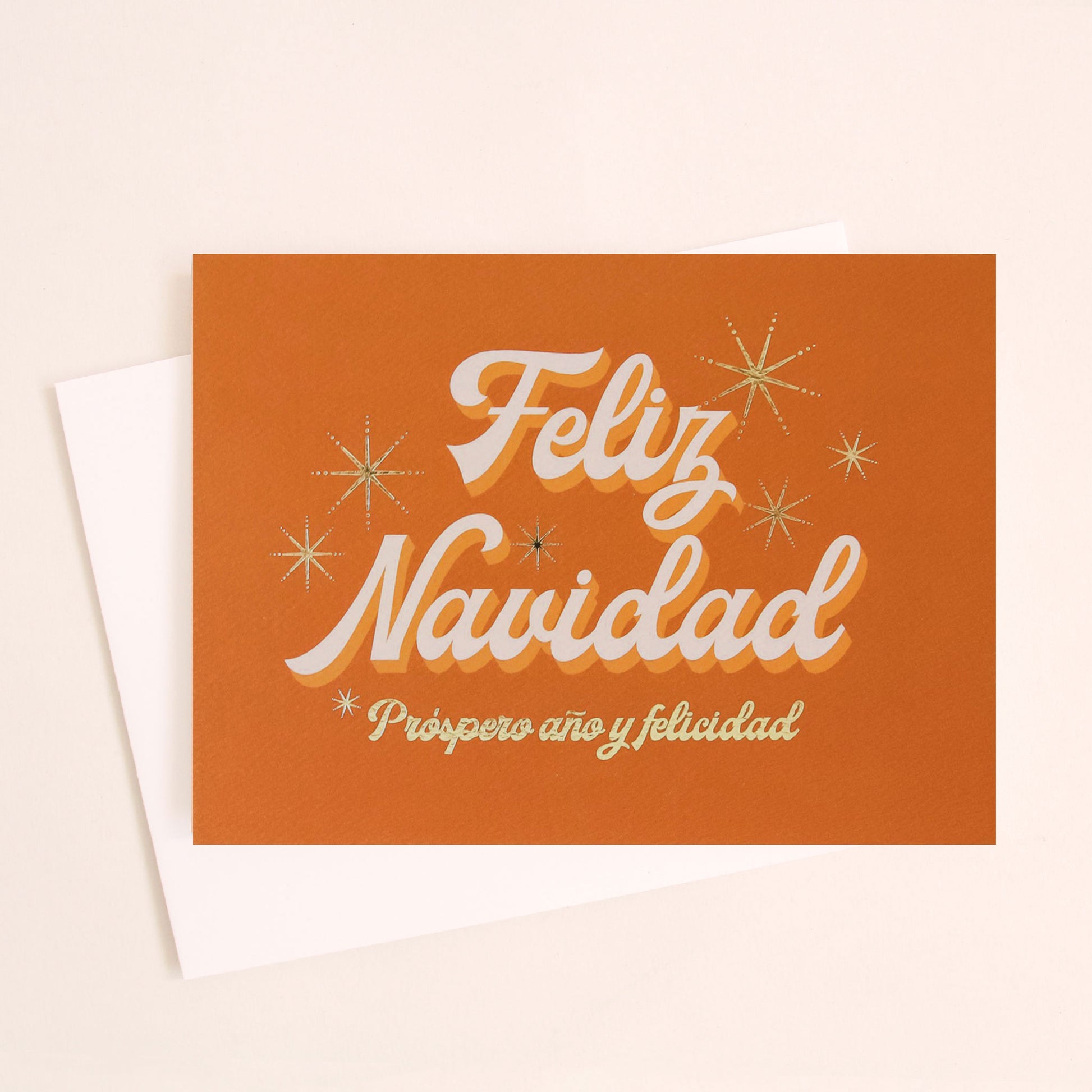 Burnt orange greeting card that reads 'Feliz Navidad' in white cursive lettering. Below reads 'prosper año y felicidad' in gold foil lettering. Gold foil stars twinkle behind the text. 