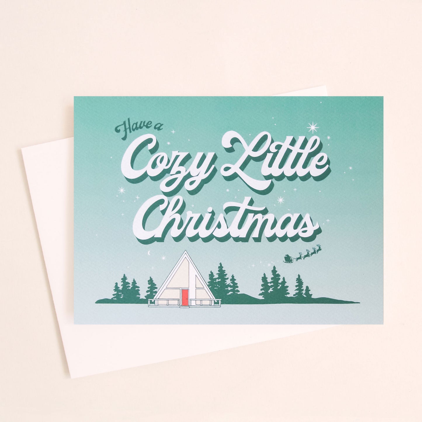 Aqua and white ombre greeting card that reads 'have a cozy little Christmas'. Below the text is a small white tent in a forest scene. A small Santa sleigh and reindeer fly across the sky.  The card is accompanied by a white envelope. 