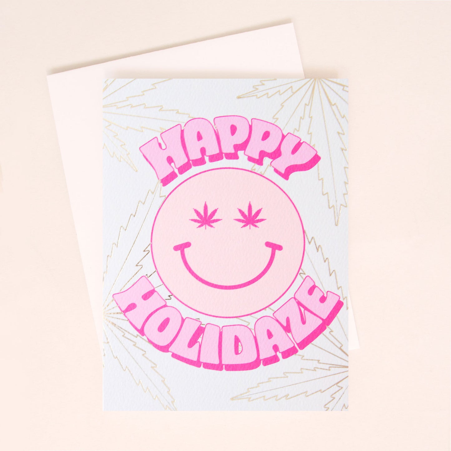 Pale card with gold foil weed leaves in the background. In the center of the card is a pale pink smiley face with magenta weed leaves for eyes. The text 'Happy Holidaze' curves around the smiley face. The card is accompanied by a white envelope.