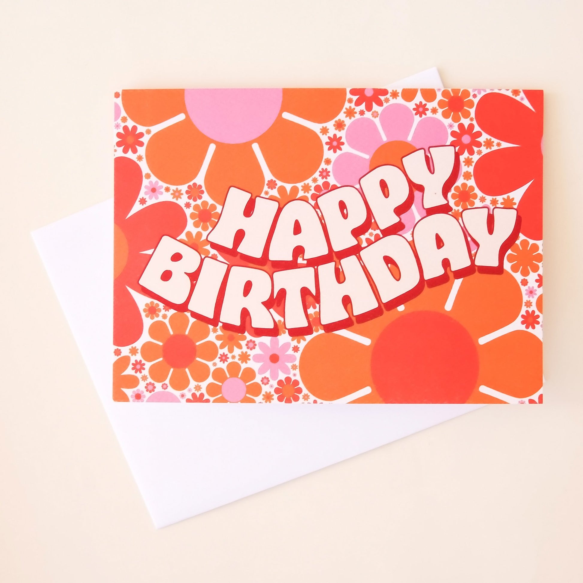 Card covered in orange and purple retro flower print that reads 'Happy Birthday' in curved bubble lettering. The card is accompanied by a white envelope.  