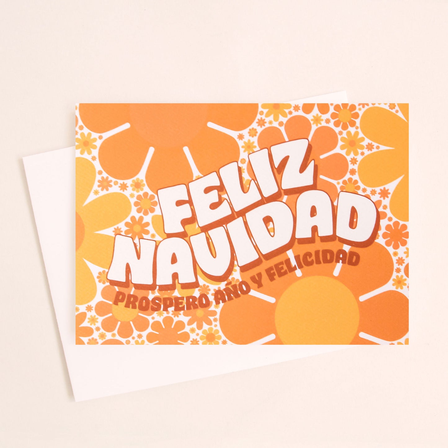 Greeting card filled with yellow and orange retro flower print. The card reads 'Feliz Navidad' in white curved bubble letters. Below reads 'prosper año y felicidad' in rust colored lettering. The card is accompanied by a solid white envelope. 