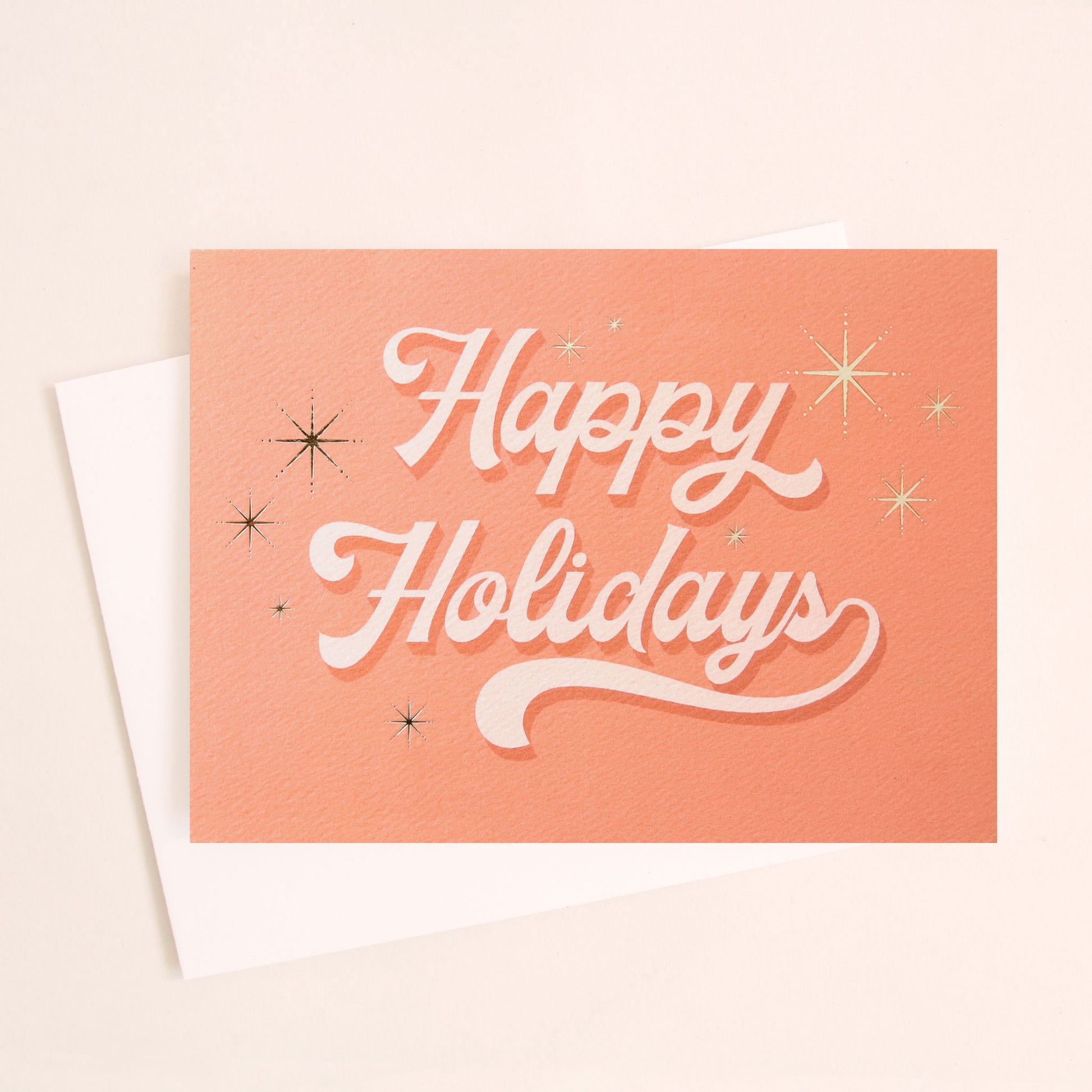 Pink greeting card with white cursive text that reads happy holidays surrounded by six gold foil starbursts. The card is accompanied by a solid white envelope.