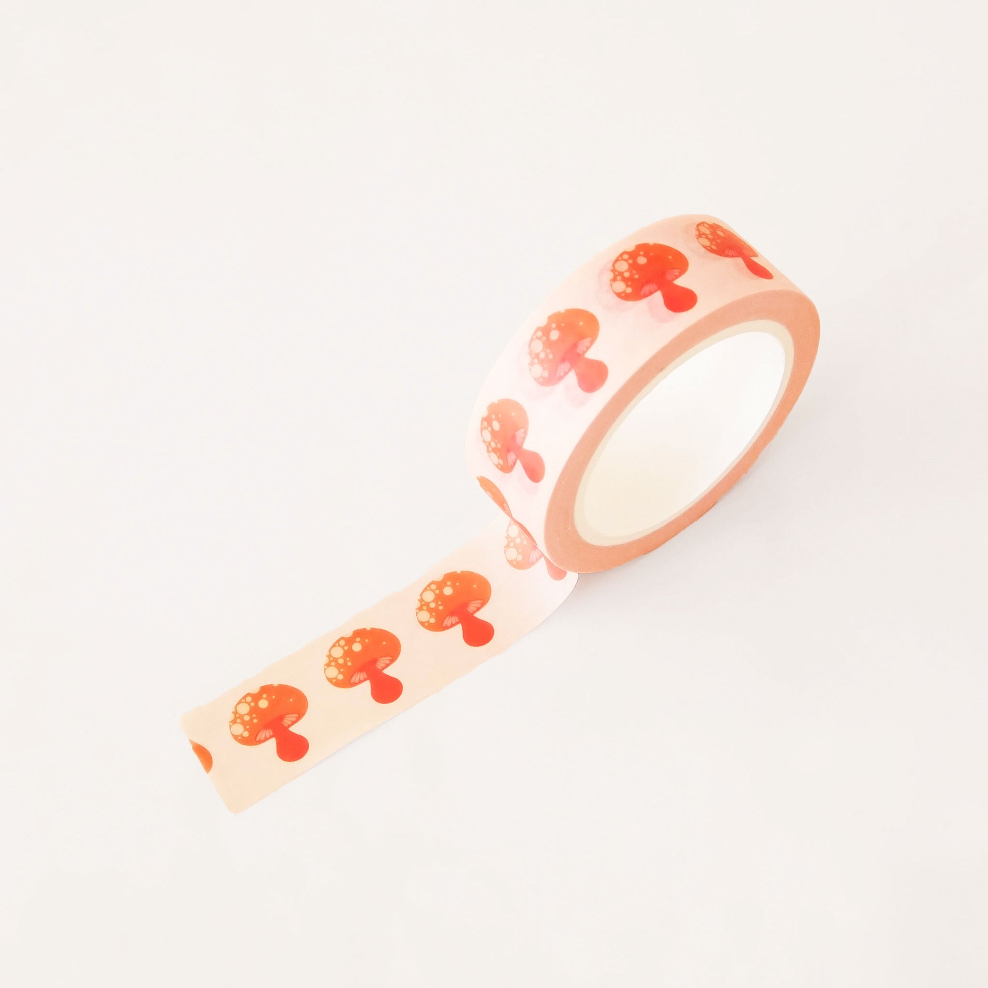 Roll of peach washi tape covered in red classic mushrooms. The roll is position upright, partially unrolled. 
