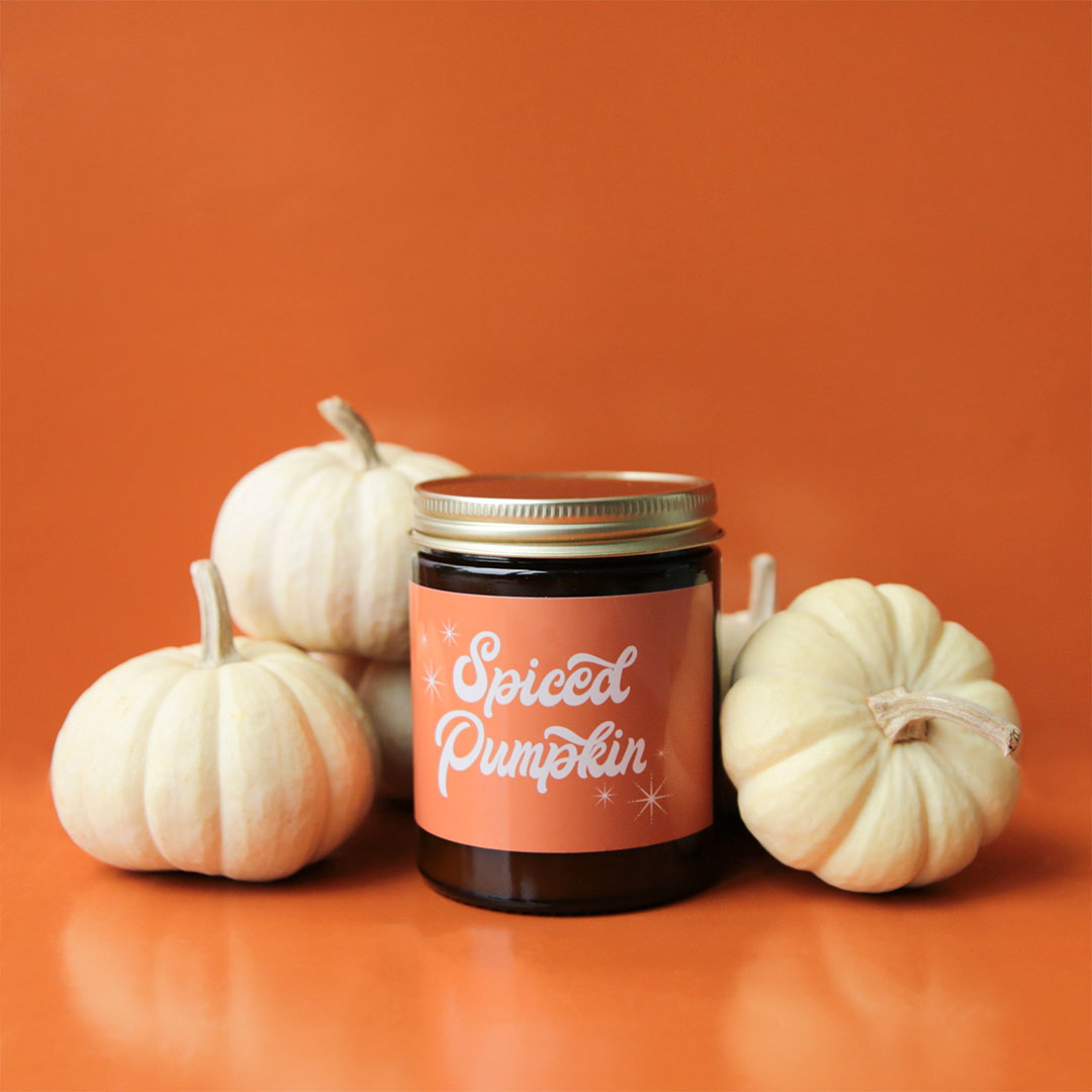 Against an orange background is a dark amber glass candle jar with an orange label that reads, "Spiced Pumpkin" in white cursive font. Also photographed are 5 white pumpkins staged beside the candle. Pumpkins not included.