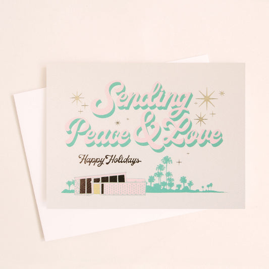 Cream card reading 'Sending Peace & Love' in soft pink lettering with seafood shadowing. Below reads 'Happy Holidays' in black cursive lettering. Below is a coastal scene of a pink beach house and sea foam palms.