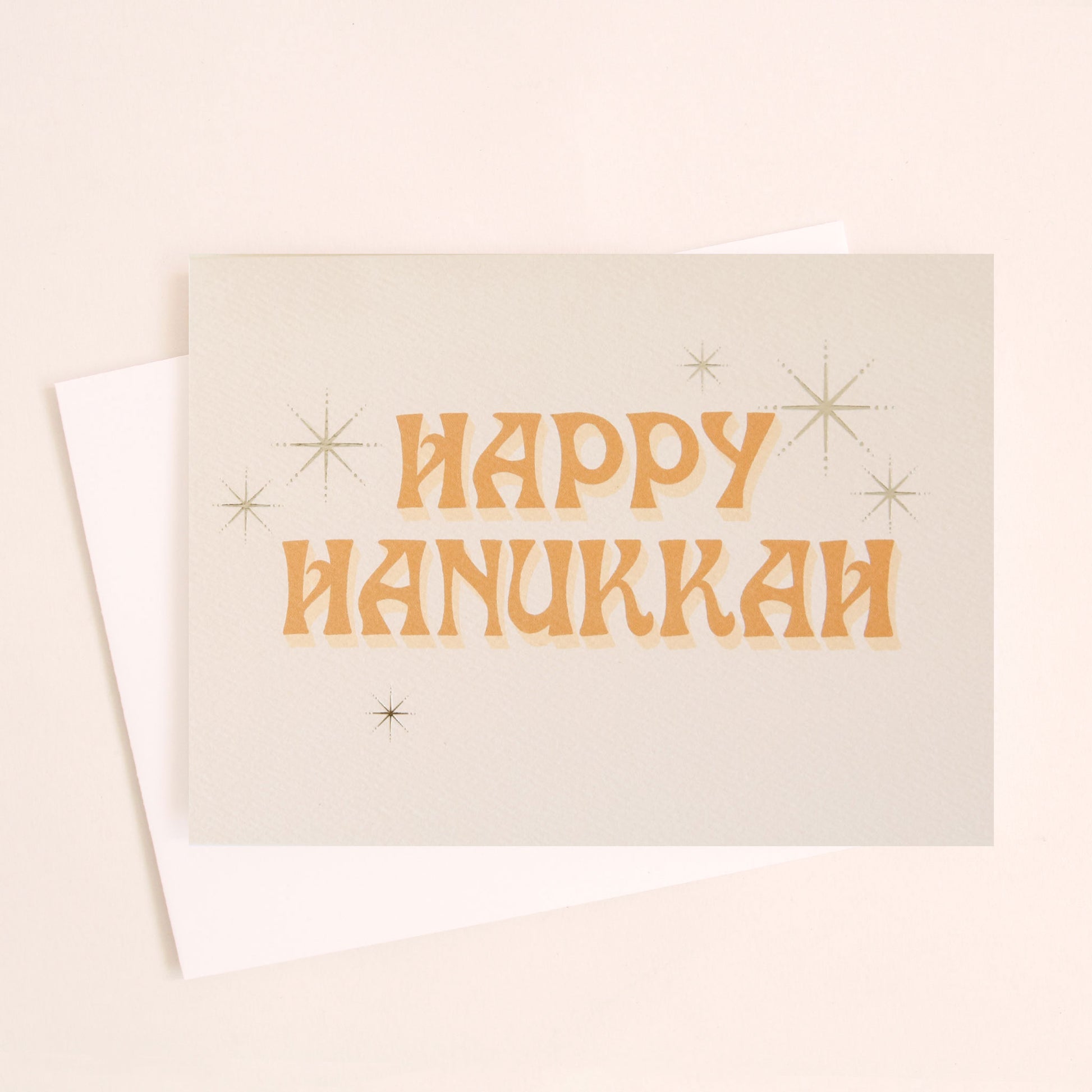 Ivory card that reads 'Happy Hanukkah' in retro orange lettering. The text is accented with gold foil stars twinkling around. 