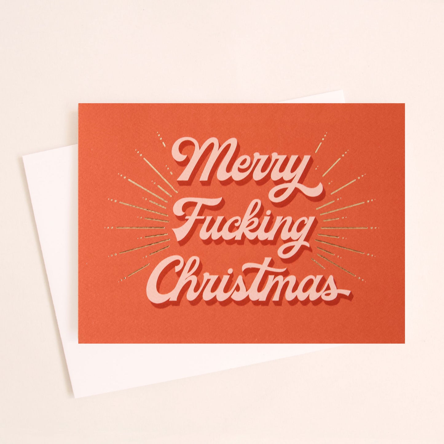 Red card that reads 'Merry Fucking Christmas' in soft pink lettering with dark red shadowing. A gold foil burst detailing beams from the text.