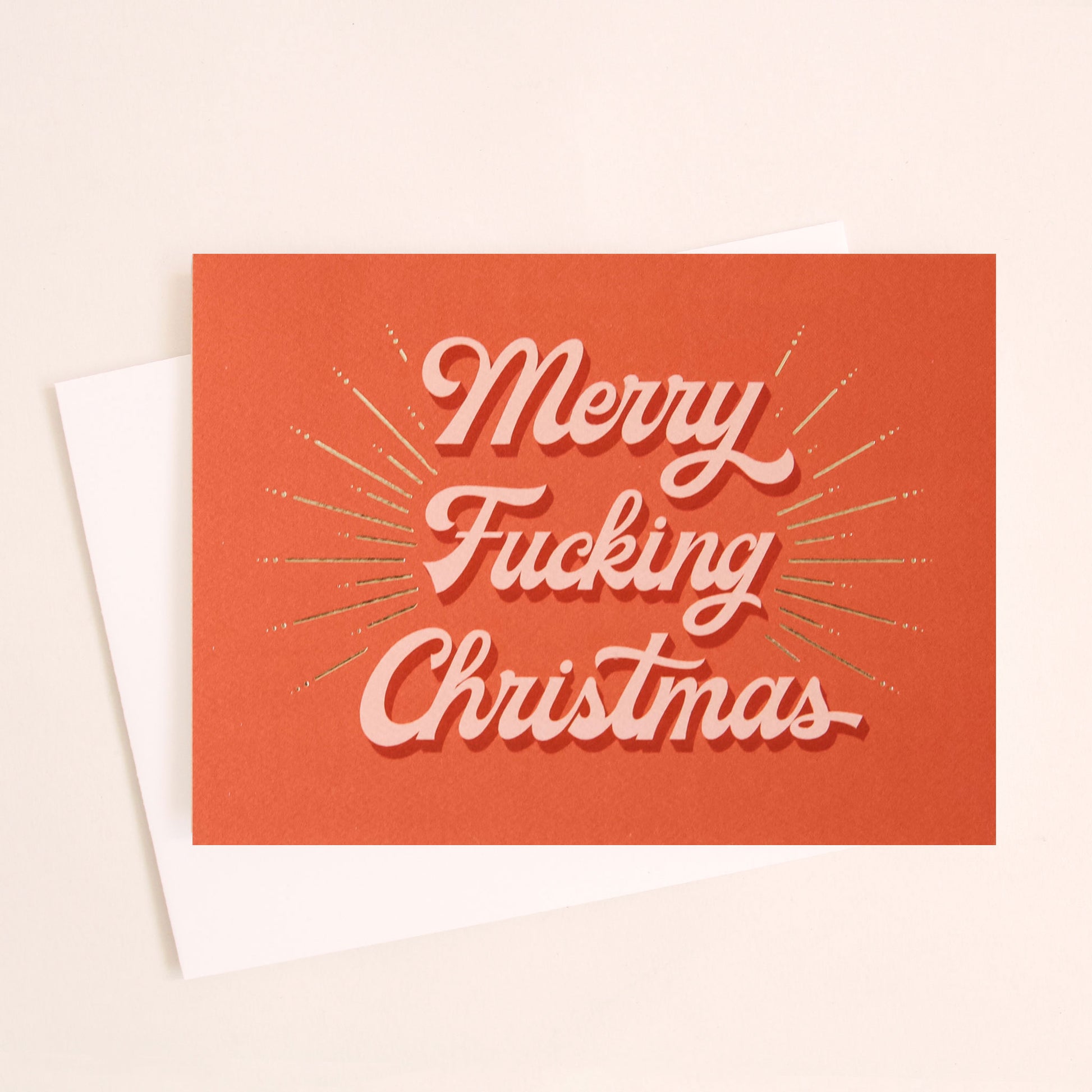 Red card that reads 'Merry Fucking Christmas' in soft pink lettering with dark red shadowing. A gold foil burst detailing beams from the text.