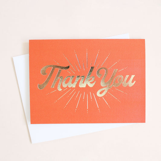 Tangerine orange card reading 'Thank You' in gold foil. Accent marks beam from the text. The card is accompanied by a solid white envelope