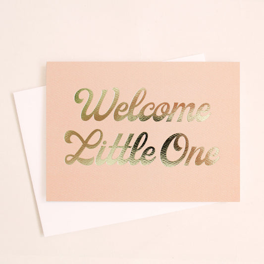 This card is a cloud peach color and reads 'Welcome Little One' in gold foil cursive lettering. The text takes up majority of the card. The card is accompanied by a solid white envelope.