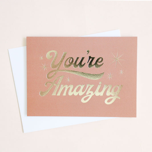 This soft pink card reads in 'You're Amazing' in shimmering, gold foil surrounded by six twinkling stars. This card is accompanied by a white envelope.
