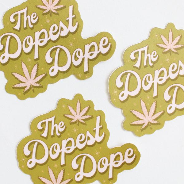 Zoomed in view of three olive green stickers that read 'The Dopest Dope' in pastel pink surrounded by two pink marijuana leaves and twinkling detailing. 
