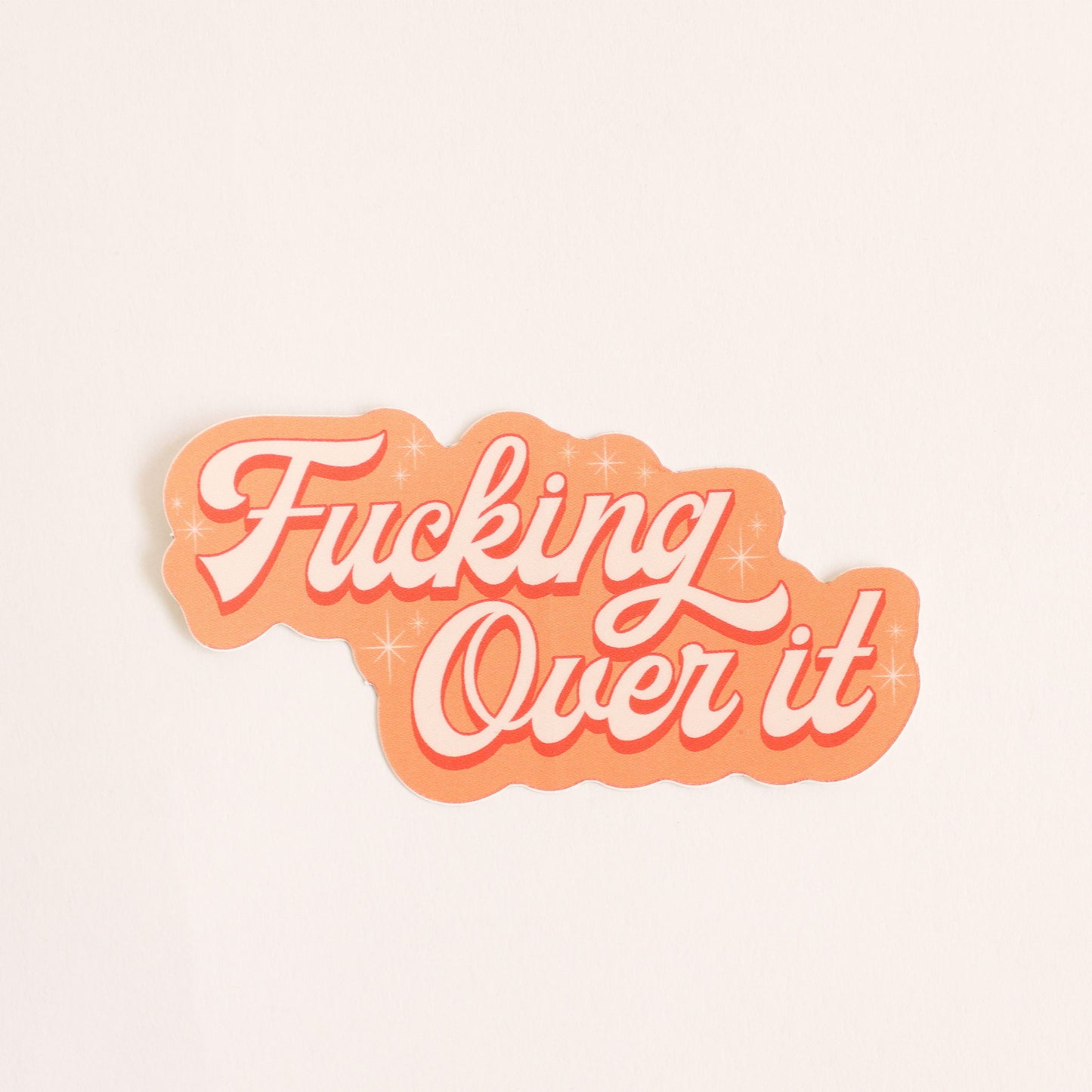 Orange toned sticker that reads 'fucking over it' in light cursive lettering. Small twinkle detailing accents around the text. 