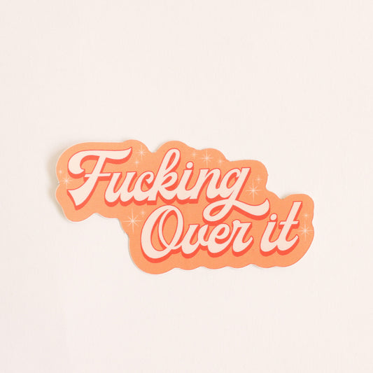 Orange toned sticker that reads 'fucking over it' in light cursive lettering. Small twinkle detailing accents around the text. 