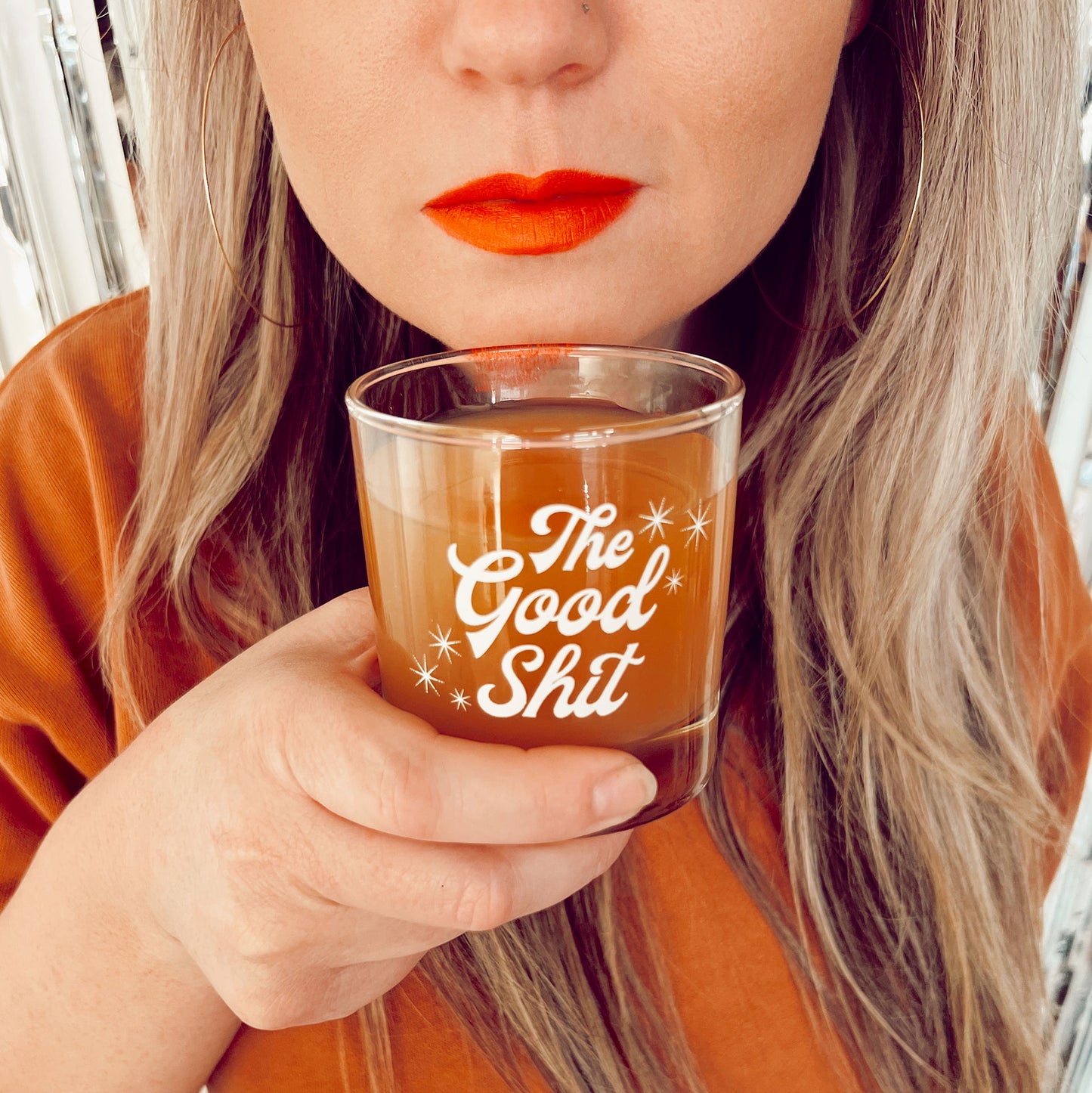 A photograph of a short glass tumbler with a thick bottom and "The Good Shit" printed across the center in white groovy cursive text.