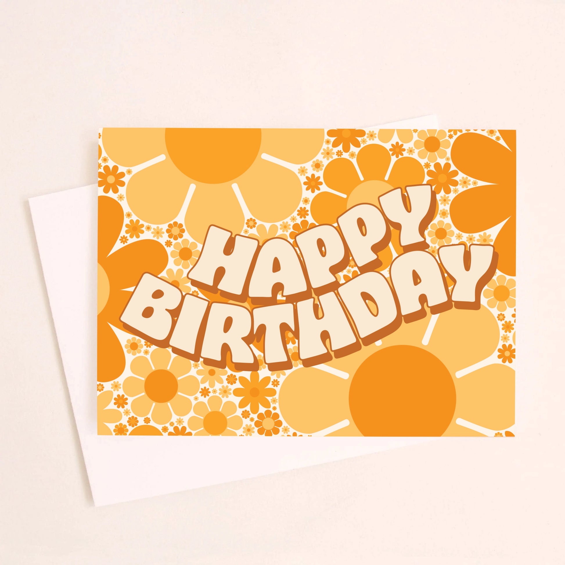 On a cream background is a card with a yellow and orange daisy print and wavy cream text in the center that reads, "Happy Birthday" alongside a white envelope. 
