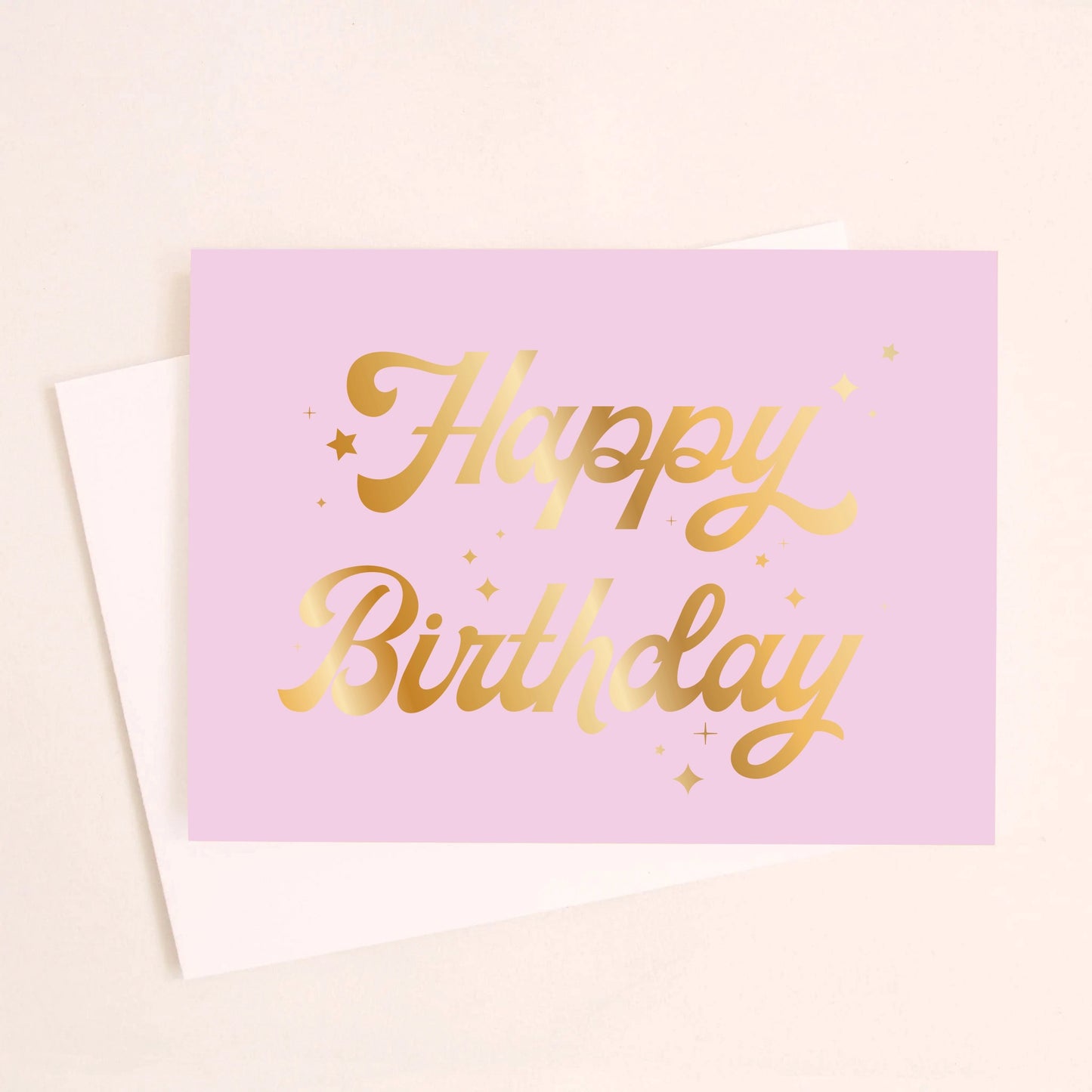 On a cream background is a lavender card with gold foiled cursive text that reads, "Happy Birthday" along with a white envelope. 
