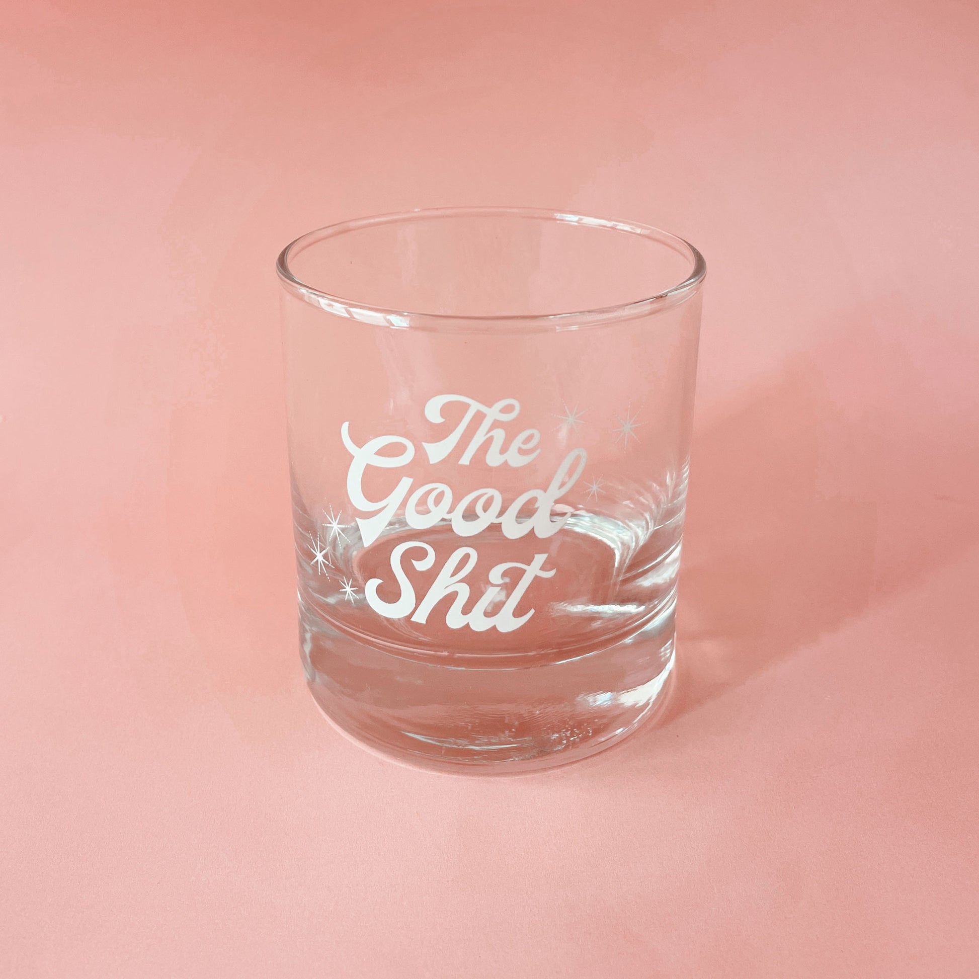 Against a peachy pink background is a photograph of a short glass tumbler with a thick bottom and "The Good Shit" printed across the center in white groovy cursive text.