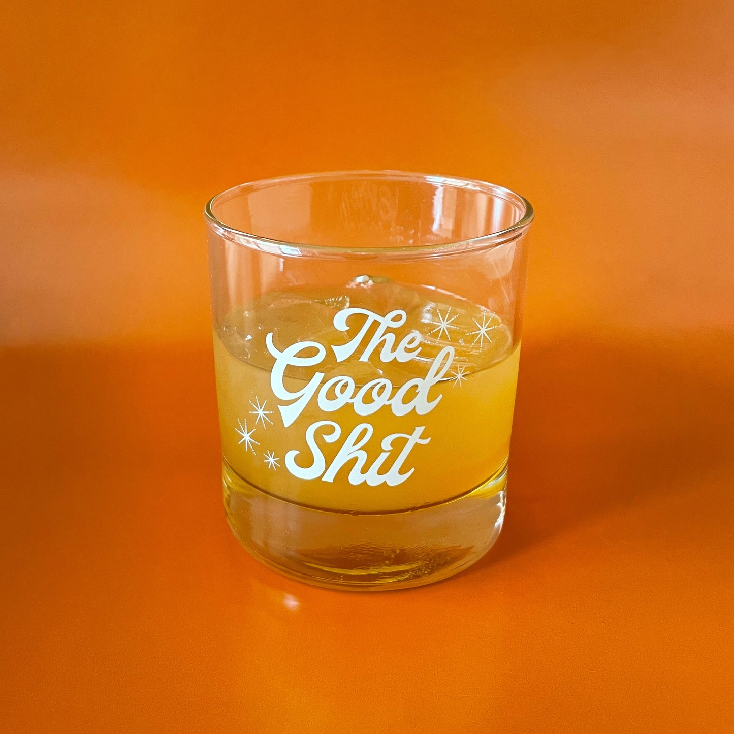Against an orange background is a photograph of a short glass tumbler with a thick bottom and "The Good Shit" printed across the center in white groovy cursive text.