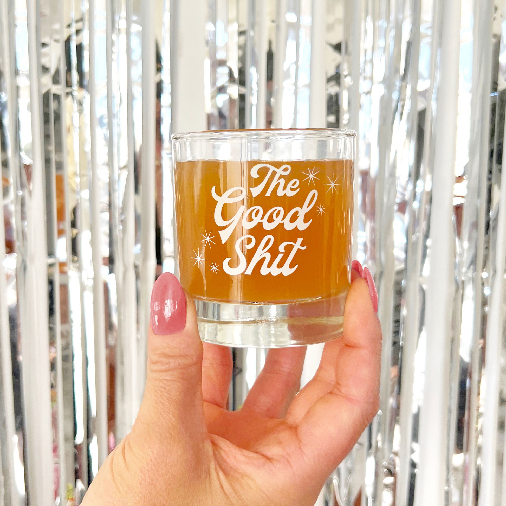 Against a silver tinsel backdrop is a photograph of a short glass tumbler with a thick bottom and "The Good Shit" printed across the center in white groovy cursive text.