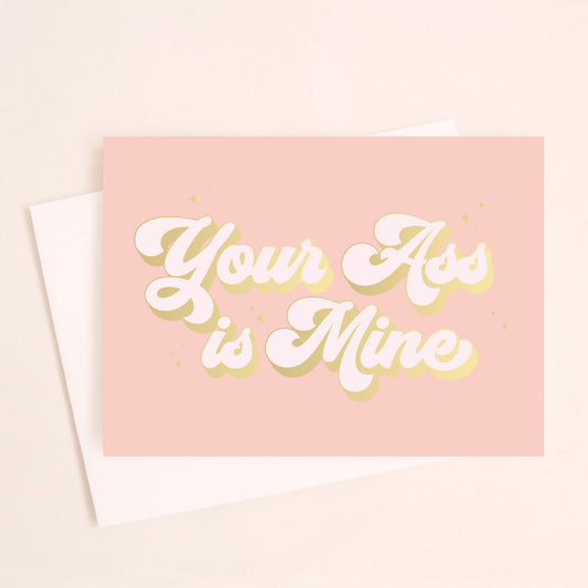 A pink card with text on the front that reads, "Your Ass is Mine" in cream text and outlined with gold foil. Also included and photographed is the coordinating white envelope.