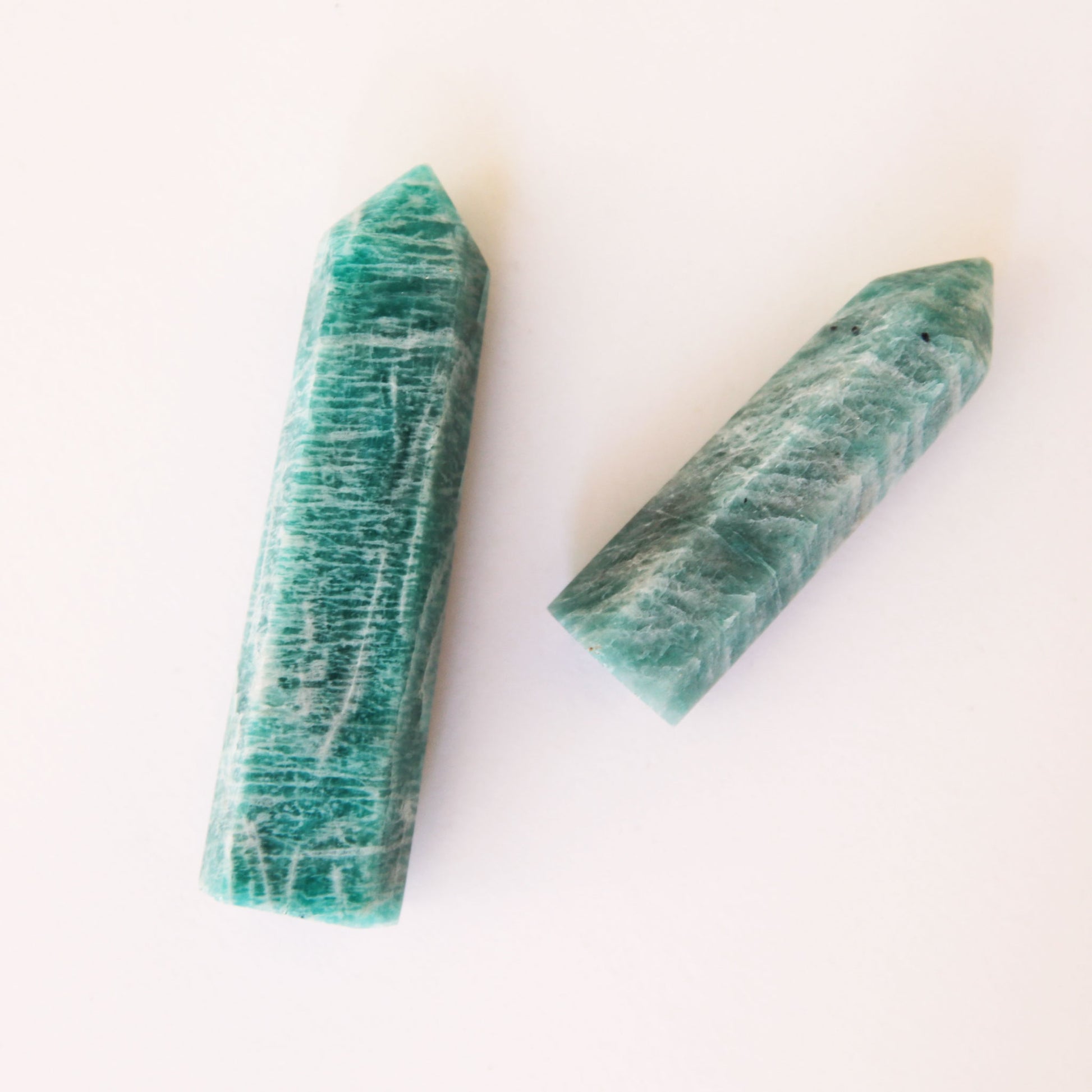 A photo of two amazonite crystals, teal blue in color with white accents. The two sizes represent the two sizes available for purchase. Each crystal is sold separately. These amazonite crystals are in the shape of a point and are flat on the bottom to allow them to be freestanding.